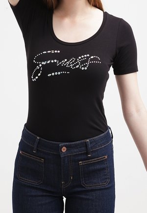 Kvinde iført en sort tætsiddende T-shirt med et "Guess"-logo, der er udsmykket med rhinsten, og mørkeblå jeans med forlommer.