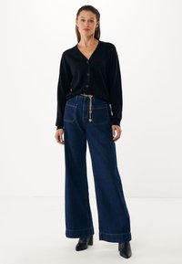 Cardigan nero con bottoni, pantaloni denim blu scuro a gamba larga e stivali neri con tacco. Dettaglio catena dorata sui pantaloni. Texture liscia.