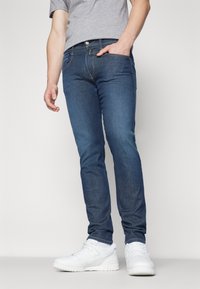 Vaqueros de mezclilla azul oscuro con corte ajustado. Diseño de cinco bolsillos, cierre con botón de metal y detalles de costura sutiles. Combinados con zapatillas blancas.