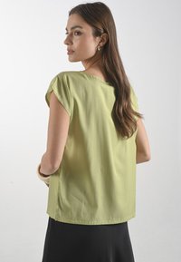 Blůza s krátkým rukávem v světle zelené barvě, s volným střihem a hladkou texturou. Zadní pohled ukazuje jednoduchý výstřih a rovný lem.