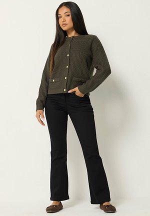 Donna con i capelli lunghi e scuri che indossa un cardigan verde scuro abbottonato, pantaloni neri e ballerine marroni, in piedi davanti a uno sfondo chiaro.