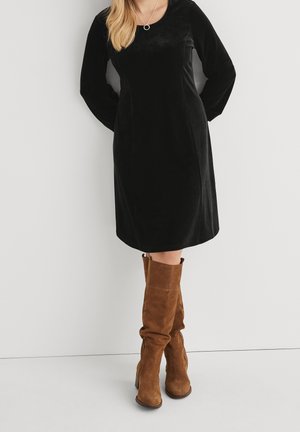 Žena nosí černé sametové šaty po kolena s dlouhými rukávy, hnědé semišové kozačky ke kolenům a jednoduchý náhrdelník s přívěskem.