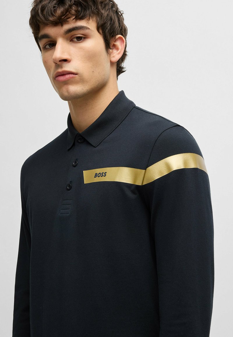 Schwarzes Polohemd aus glattem Stoff, mit einem goldenen Streifen und dem "BOSS"-Logo auf der Brust. Klassischer Kragen und Knopfleiste.