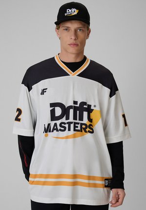 Młody mężczyzna w białej koszulce Drift Masters z czarnymi i żółtymi akcentami oraz czarną czapeczką Drift Masters, zwrócony przodem na szarym tle.