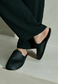 Black leather slip-on shoes with a smooth texture and rounded toe, set against a neutral background. Foot partially visible.
