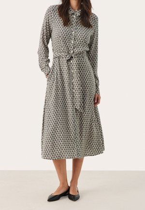 Vrouw met een midi-jurk met lange mouwen en knopen, een patroonriem en zwarte platte schoenen, staand tegen een effen achtergrond.