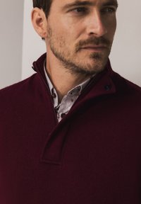 Bordeauxroter Pullover mit Viertel-Reißverschluss aus weichem Stoff, mit einem strukturierten Kragen und einem darunter getragenen Hemd mit dezentem Muster.