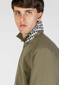 Giacca verde oliva con chiusura a bottoni, con colletto a scacchi in nero, bianco e blu. Texture liscia, vestibilità sartoriale.