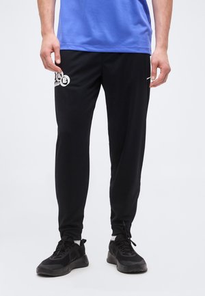 Férfi fekete Nike sportnadrágban, logóval és fekete tornacipőben, karjai az oldalán lazán lógva áll.