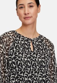 Schwarz-weiße gemusterte Bluse mit einem Schlitz-Ausschnitt, durchsichtigen Ärmeln und einem weichen, strukturierten Stoff mit einem blattähnlichen Design.