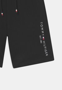 Pantaloni corti neri con tasche frontali e cordino, caratterizzati da testo e logo verticali bianchi "Tommy Hilfiger Est. 1985" sulla gamba destra.