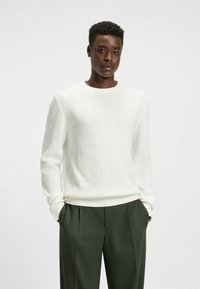 Weißer Strickpullover mit einem strukturierten Zickzackmuster, rundem Ausschnitt und weitem Bund, kombiniert mit grünen, plissierten Hosen.
