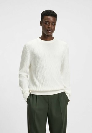 Weißer Strickpullover mit einem strukturierten Zickzackmuster, rundem Ausschnitt und weitem Bund, kombiniert mit grünen, plissierten Hosen.