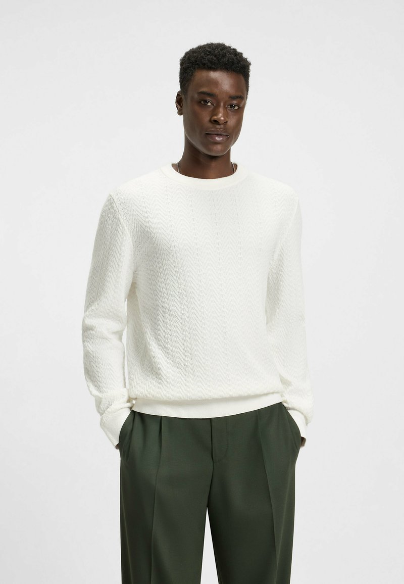 Weißer Strickpullover mit einem strukturierten Zickzackmuster, rundem Ausschnitt und weitem Bund, kombiniert mit grünen, plissierten Hosen.