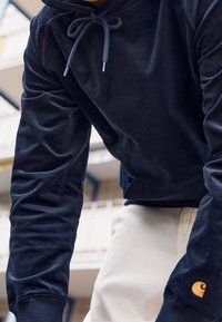 Navy Kapuzenpullover mit Kordelzug, gerippten Bündchen und strukturiertem Finish. Verfügt über ein kontrastierendes goldenes Logo auf dem linken Ärmel.