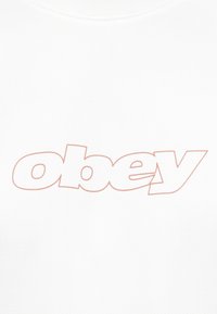 Λέξη 'obey' με διακριτό περίγραμμα σε ανοιχτό καφέ χρώμα σε απλό λευκό υφασμάτινο φόντο, κεντραρισμένη και έντονη με συνδεδεμένα γράμματα.