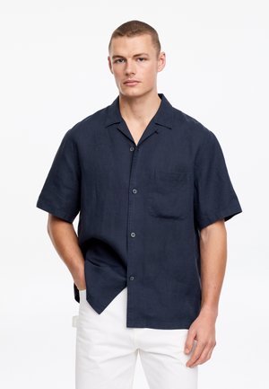 Jeune homme portant une chemise bleu marine à manches courtes avec une poche poitrine et un pantalon blanc, debout avec une main dans la poche.