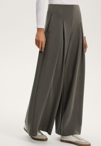Szerokie spodnie w ciemnoszarym kolorze, z przodu z zakładkami i gładką fakturą. W zestawieniu z jasnymi trampkami, co tworzy swobodny styl.
