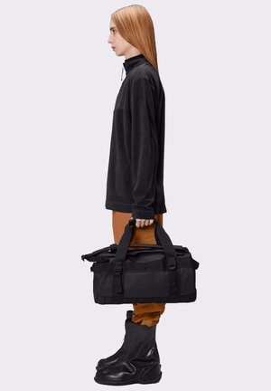 Borsa a tenuta doppia nera in tessuto testurizzato, con maniglie laterali, tenuta da una persona che indossa un maglione di pile nero e pantaloni marroni, con stivali neri.