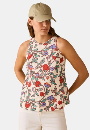 Donna con cappellino beige, canotta senza maniche con motivi floreali rossi e viola, e pantaloni chiari, in piedi con le mani dietro la schiena.