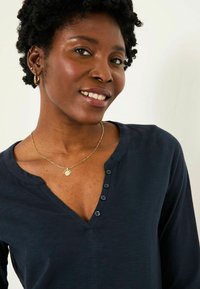 Navy long-sleeve top with a V-neckline and five small buttons. Gold chain necklace with a circular pendant accentuates the neckline.