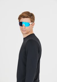 Zonnebril met grote blauwe spiegellenzen, een wit frame en een sportief design, gedragen door een model in een strakke zwarte longsleeve.