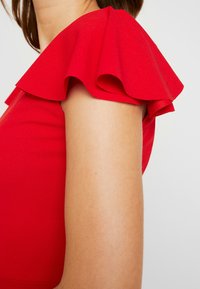 Czerwony strój z szerokim, falbaniastym rękawem. Tkanina wydaje się gładka, o dopasowanym kroju podkreślającym ramię.