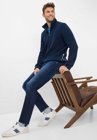 Pile blu navy con colletto alto, abbinato a jeans blu scuro e sneakers bianche con dettagli blu navy, seduto su una sedia di legno.