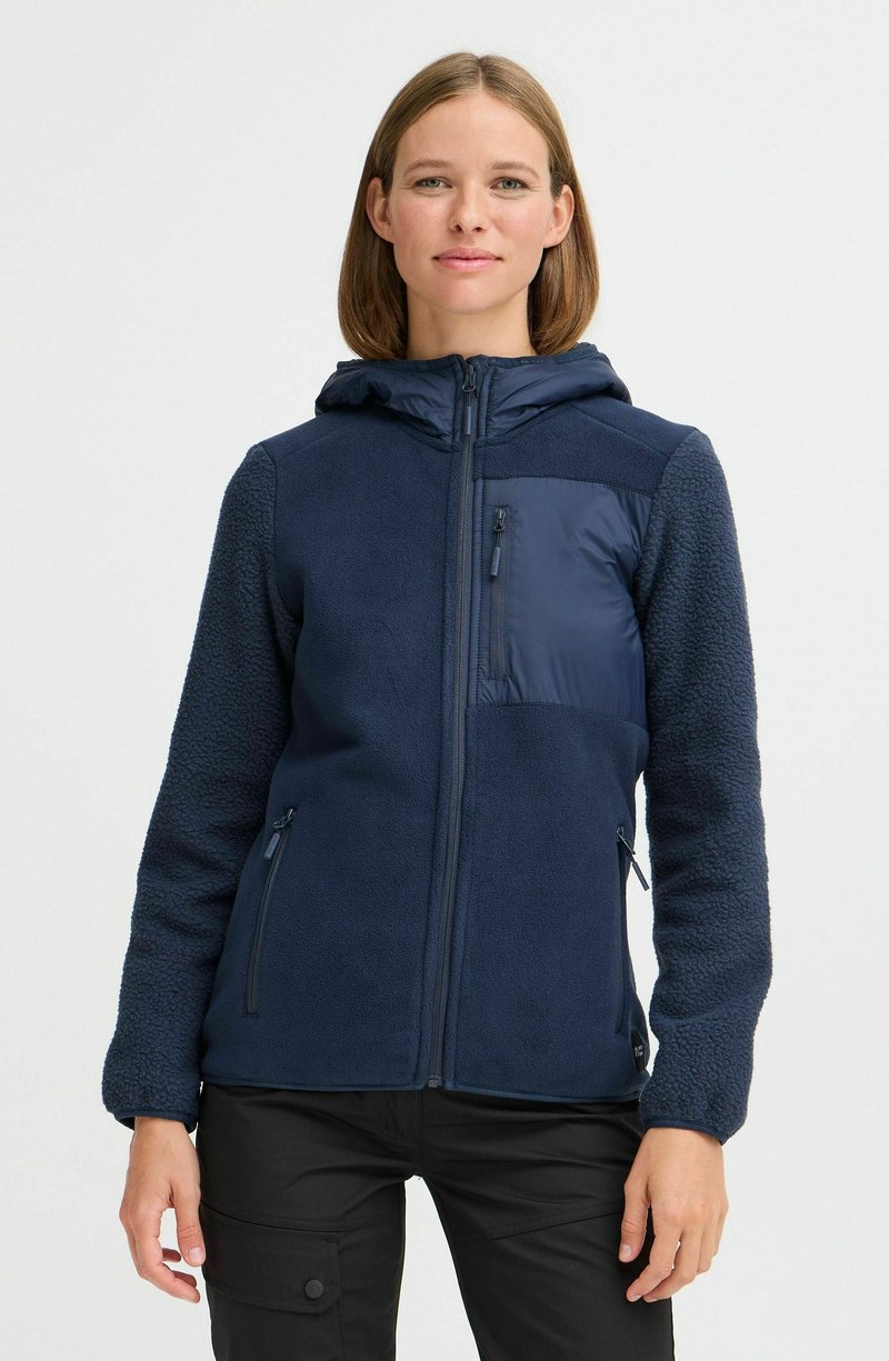 Veste en polaire bleu marine avec fermeture éclair à l'avant, capuche et diverses textures. Elle comprend un panneau lisse matelassé et des poches zippées.