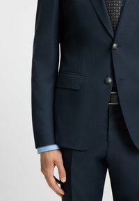 dunkelblau strukturierte Anzugjacke mit einem einzigen Knopfverschluss, zwei Taschen und einem hellblauen Hemdärmel, der unter dem Ärmel hervorblitzt.