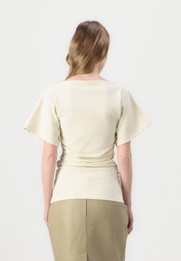 Haut beige clair avec des manches courtes volantes, présentant une silhouette ajustée et des détails froncés à la taille, associé à une jupe kaki.