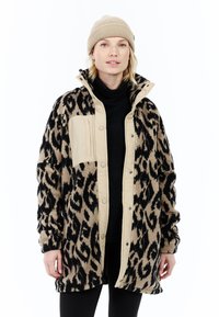 Žena u bež kapici i flis jakni s leopard uzorkom preko crne dolčevite, stoji protiv bijele pozadine.
