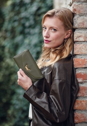 Green leather wallet with a textured finish, featuring a subtle curved design and gold hardware logo. Model holds it against a brick wall.
