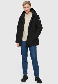 Vrouw staat in een zwart hooded jack, beige trui, blauwe jeans en zwarte enkellaarsjes tegen een effen lichte achtergrond.