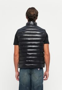 Bărbat îmbrăcat cu o vestă puffer neagră peste un tricou negru cu mânecă scurtă și blugi, întors cu spatele, pe un fundal alb simplu.