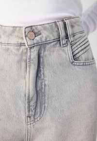 Lys grå denimjeans har en knappelukking, subtile falminger og broderte detaljer på lommene. Teksturen er glatt med en svak glans.