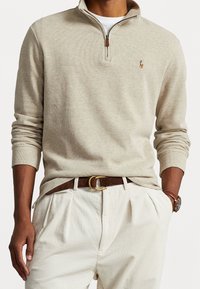 Beige zip-up tröja med ribbad textur och en brun läderdragkedja, tillsammans med ljusa, veckade kordbyxor och ett brunt bälte.