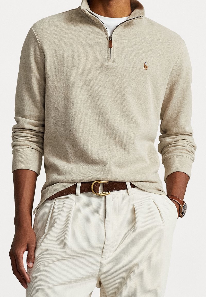 Beige zip-up tröja med ribbad textur och en brun läderdragkedja, tillsammans med ljusa, veckade kordbyxor och ett brunt bälte.