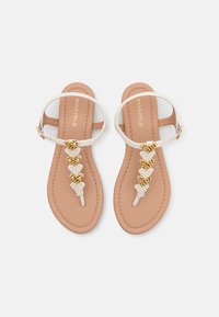Sandales plates blanches avec un design T-strap ornées d'accents en cœur en or et perles, montées sur une semelle rembourrée beige.