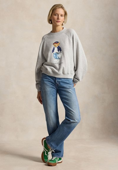 Γυναίκα που στέκεται φορώντας γκρι φούτερ με σχέδιο αρκουδάκι, μπλε τζιν και πράσινα αθλητικά παπούτσια σε ουδέτερο φόντο.