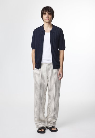 Marine tekstureret kortærmet skjorte, hvid tanktop, stribede beige brede bukser og sorte sandaler, stående mod en ensfarvet baggrund.