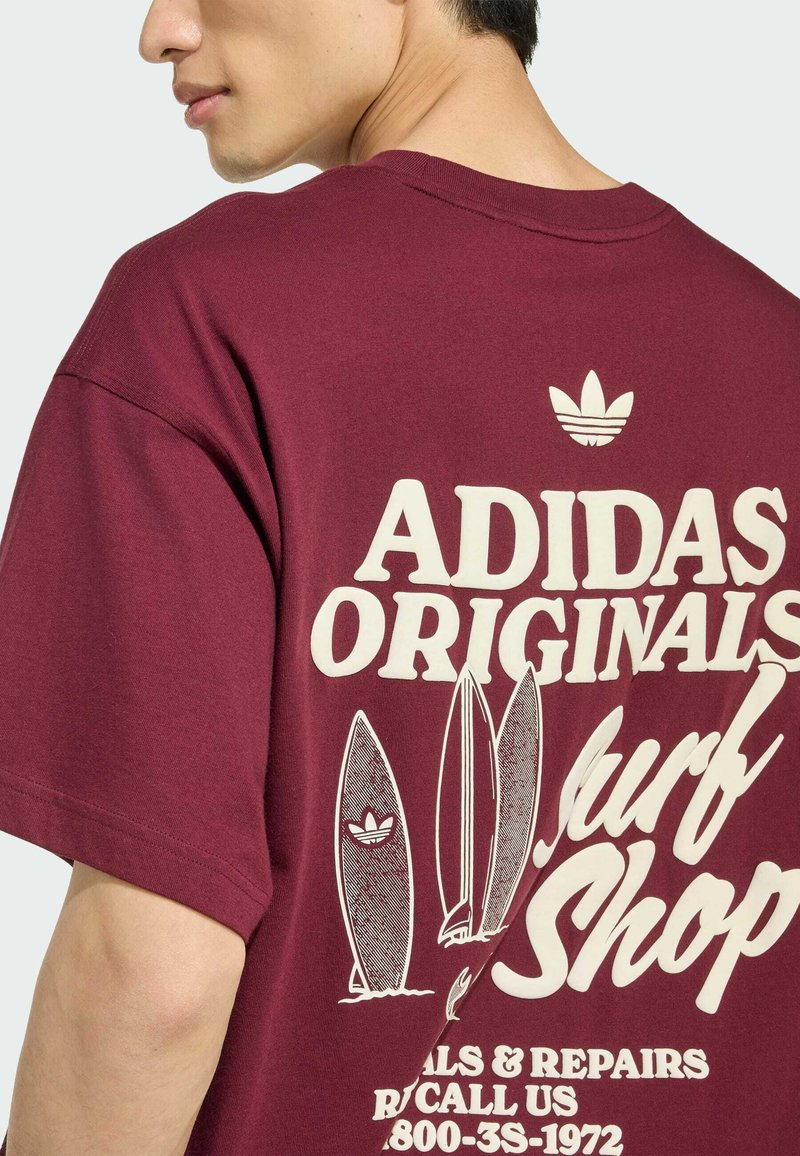 Mann trägt ein bordeauxrotes T-Shirt mit weißem "ADIDAS ORIGINALS Surf Shop"-Schriftzug und Surfbrett-Grafiken auf dem Rücken.