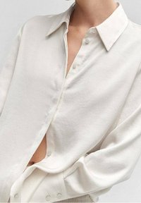 White button-up shirt made of a soft, smooth fabric. Features a classic collar, long sleeves, and a slightly loose fit with a side opening.