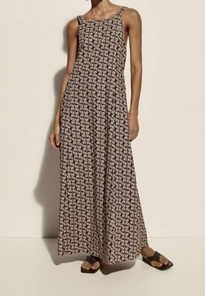 Femme portant une robe à motifs sans manches, longue jusqu'aux chevilles, avec de larges bretelles et des sandales noires, debout devant un fond clair et uni.