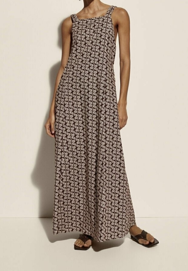 Femme portant une robe à motifs sans manches, longue jusqu'aux chevilles, avec de larges bretelles et des sandales noires, debout devant un fond clair et uni.