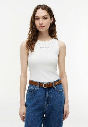 Frau trägt weißes ärmelloses Top mit dem Schriftzug „tommy jeans“ und hoch taillierte blaue Jeans mit braunem Gürtel vor einfarbigem Hintergrund.