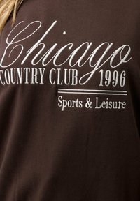 Ruskea puuvillapaita, jossa on valkoisia brodeerattuja tekstejä: "Chicago Country Club 1996" ja "Sports & Leisure" klassisella fonttityylillä.