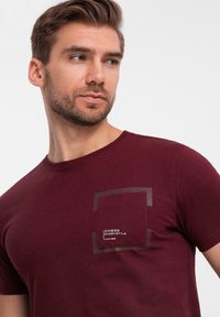 Mörkröd bomulls t-shirt med en fyrkantig grafisk ficka på vänster bröst, med vit text och en kontrasterande mörkare rektangulär kant.