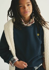 Sudadera azul marino con un logo amarillo, superpuesta a una camisa de cuadros. Suéter blanco de punto trenzado colgado sobre los hombros. Tela texturizada.