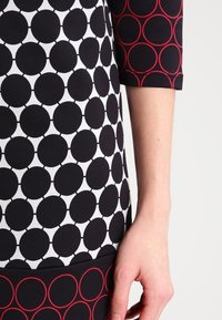 Mustavalkoinen mekko, jossa on pyöreä kuvio, teksturoitu kangas ja punaiset pyöreät yksityiskohdat hihoissa. Slim-fit-malli.